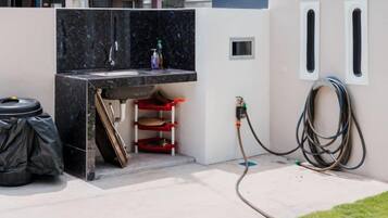 A small outdoor kitchen area with a sink, a red stool, and a hose connected to a wall.