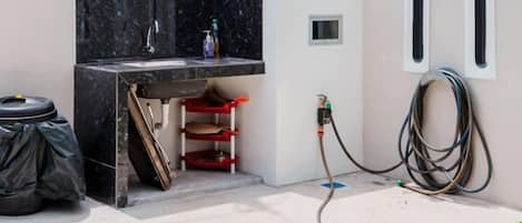 A small outdoor kitchen area with a sink, a red stool, and a hose connected to a wall.