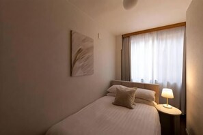 Quarto de hotel com uma cama, uma mesa de cabeceira com abajur, uma janela com cortinas e uma obra de arte na parede.