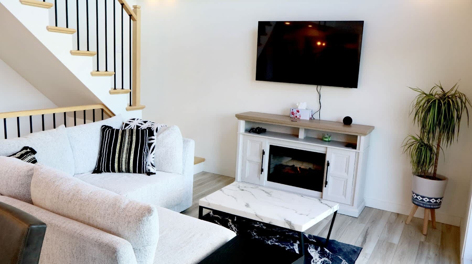 A living room with a sofa, a coffee table, a fireplace, a TV mounted on the wall, and a staircase.