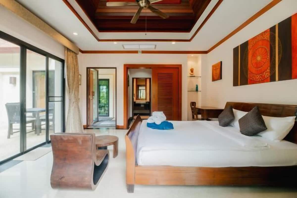 Habitación de hotel con cama, cabecera de madera, ventilador de techo, ventana grande y vistas al exterior.