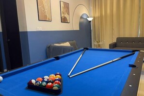 Una mesa de billar con bolas y tacos, un sofá, y una lámpara en una habitación con cortina y decoración de pared.