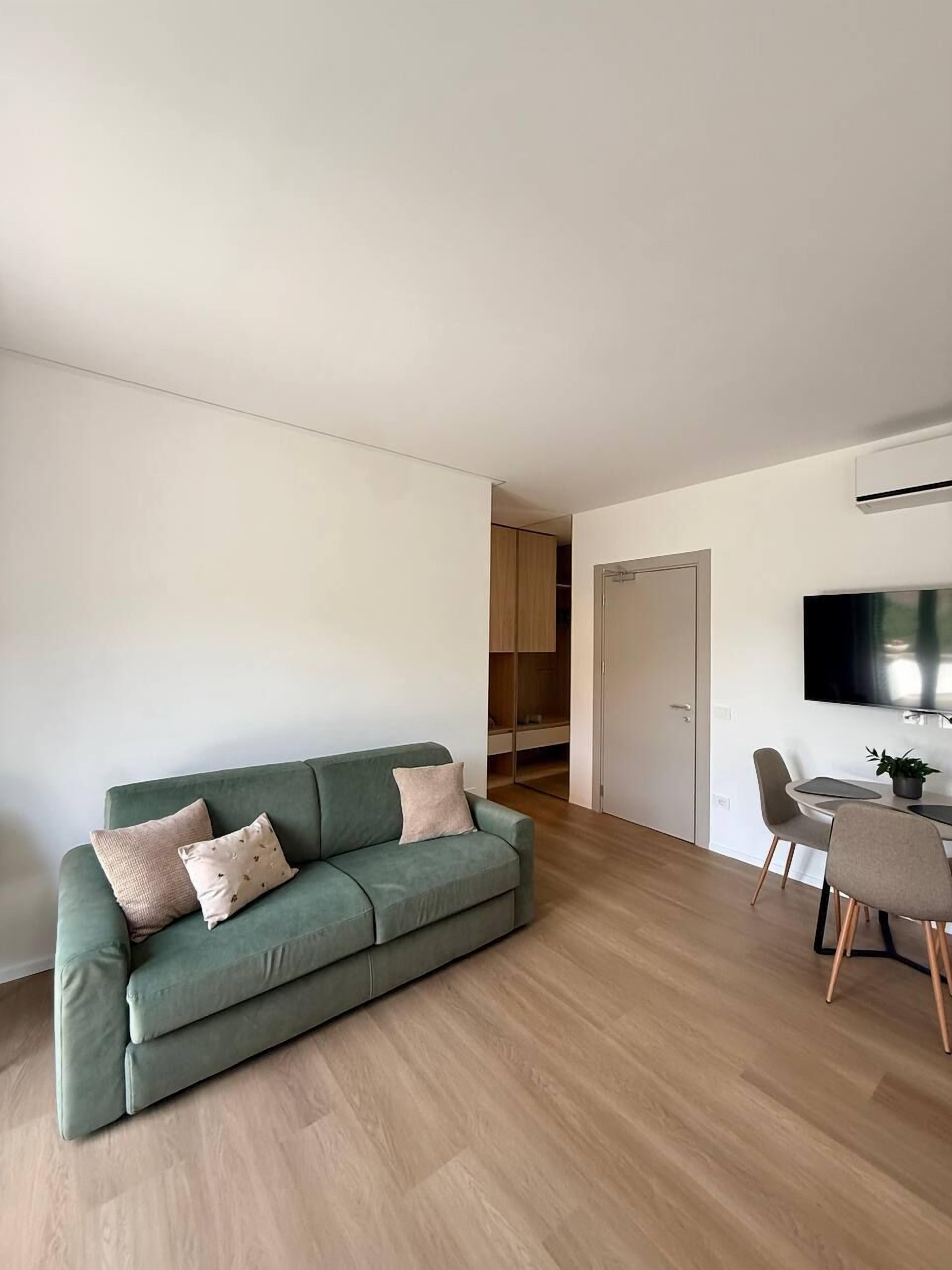 A modern living room with a green sofa, a dining area with a table and chairs, and a TV mounted on the wall.