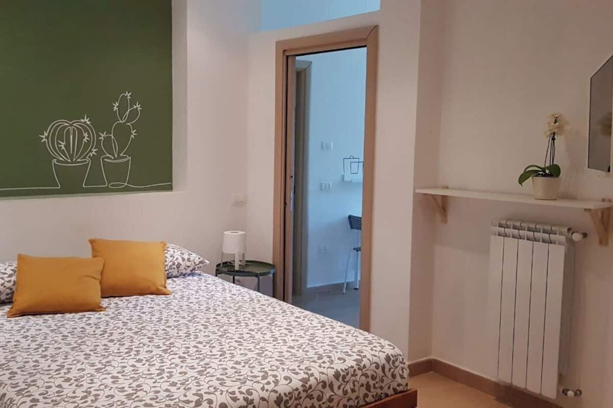 Une chambre à coucher comprenant un lit, une table de chevet, une commode, un radiateur, une fenêtre et une porte donnant sur un balcon.