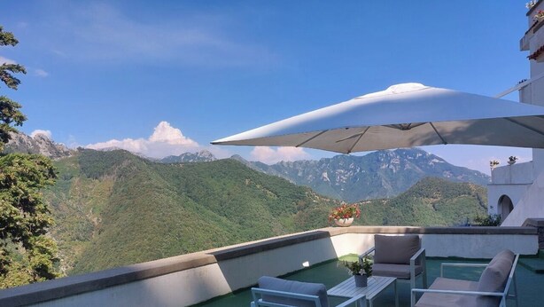 A terrace with a table and chairs, a potted plant, and a view of mountains.