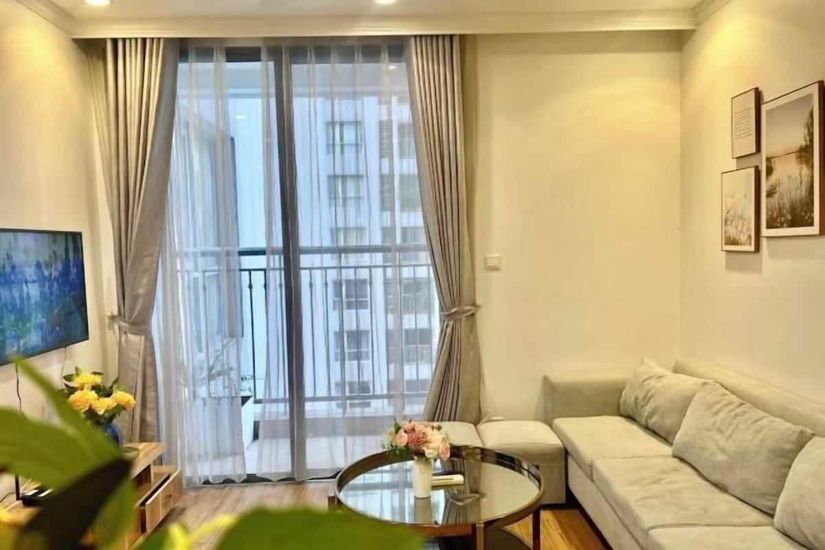 A living room with a sofa, a glass coffee table, a TV, and a large window with curtains.