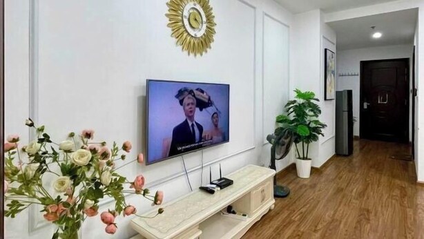 A living room with a television mounted on the wall, a decorative sunburst clock, and a potted plant. - Vinhomes Time city & Park Hill - Green Home (Hanoi)