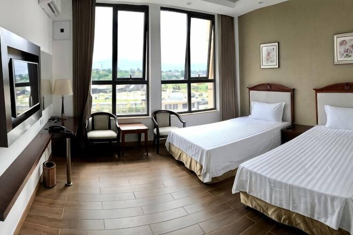 A hotel room with two beds, a desk, a chair, a TV, and large windows with a city view.