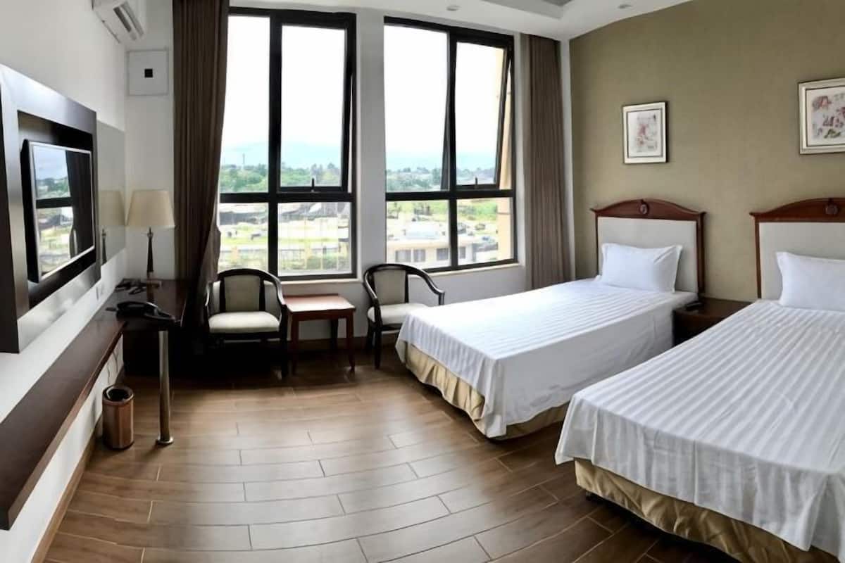 Habitación de hotel con dos camas, un escritorio, una silla, un televisor y ventanas amplias con vista a la ciudad.
