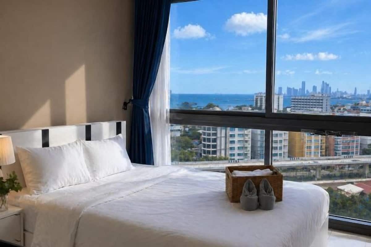 Habitación de hotel con un ventanal grande, cama con ropa de cama blanca, mesita de noche con lámpara y una cesta con zapatillas.