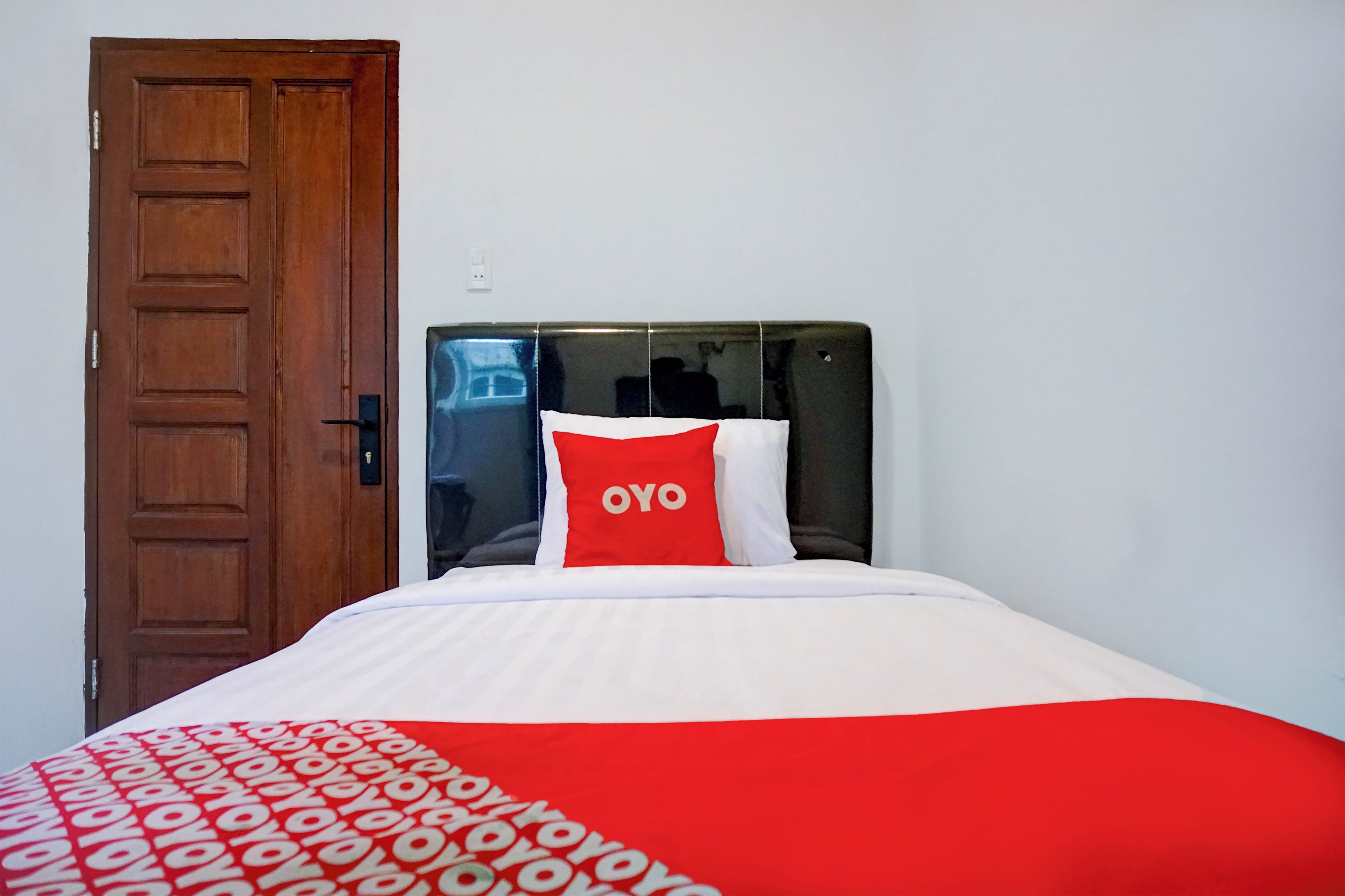 Habitación de hotel con puerta de madera, cabecero negro y cama con colcha y almohadas rojas y blancas.