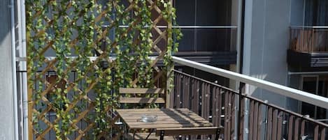 Uma varanda com mesa e cadeiras de madeira, guarda-corpo de metal e um painel treliçado com plantas trepadeiras.
