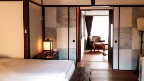 Une chambre d’hôtel avec un lit, une table de chevet avec une lampe, un banc en bois, une chaise et une vue sur l’extérieur par une porte ouverte.