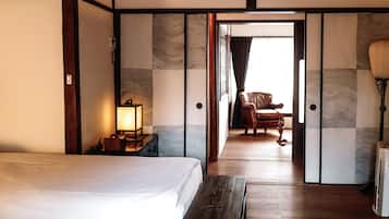 A hotel room with a bed, a nightstand with a lamp, a wooden bench, a chair, and a view through an open door.