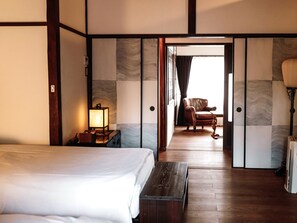 A hotel room with a bed, a nightstand with a lamp, a wooden bench, a chair, and a view through an open door. - Kyomachiya Hoshiya (Kyoto)