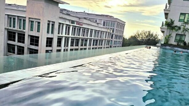 Ein Schwimmbecken mit Glasrand, neben einem Gebäude mit vielen Fenstern und einer flachen Dachterrasse.