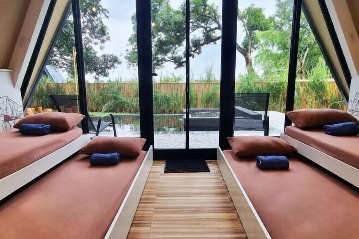 A room with two beds, a wooden floor, and a view of a garden through large windows.