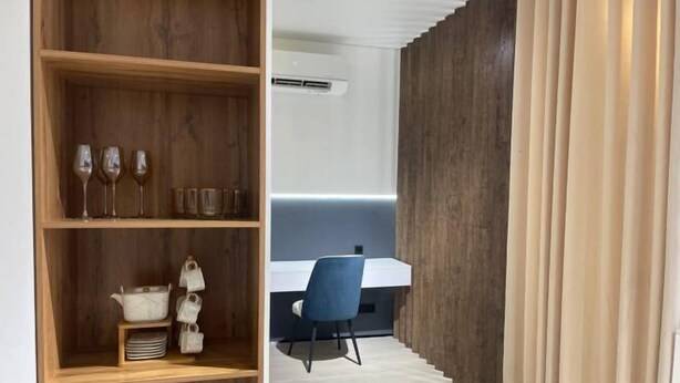 Un intérieur moderne avec une étagère en bois où sont posés des verres et des ustensiles de cuisine, un bureau blanc avec une chaise, et un mur en bois agrémenté d’un rideau.