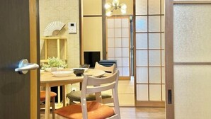 A traditional Japanese room with tatami flooring, a low table with cushions, a fan, and a sliding door leading to another room.
