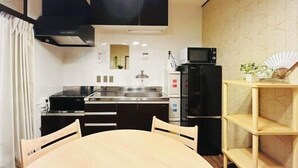 A compact kitchen with a stove, sink, microwave, and refrigerator. There is a dining table with chairs and a small shelf with a plant.