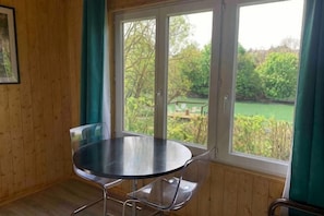 A room with a round table, a chair, a window with a view of greenery, and wooden walls. - Romantic Nature Chalet – Peaceful Escape by the Marne River (Isles-les-Villenoy)