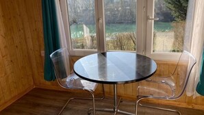 A room with a round glass table and metal chairs, wooden walls, and a view through a window.