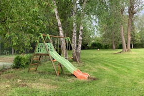 Un columpio de madera con un tobogán verde y naranja en un área cubierta de césped y con árboles.