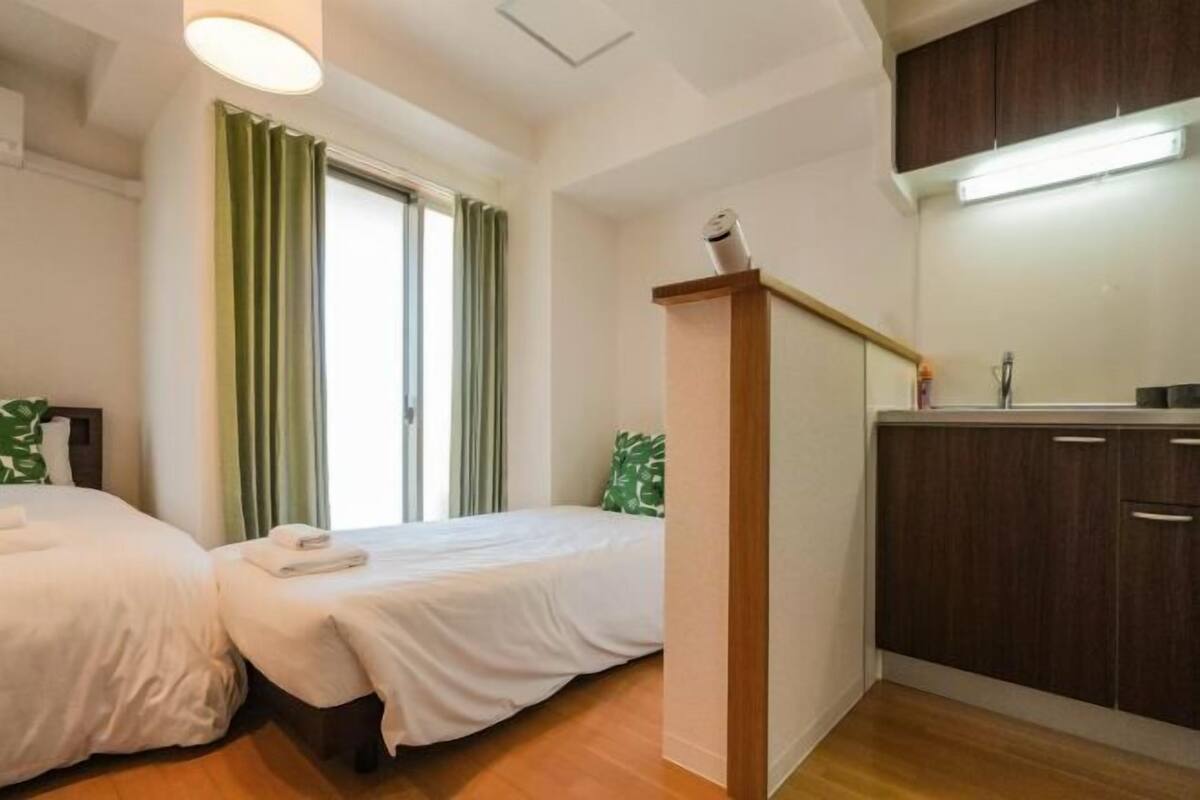 Habitación de hotel con dos camas, suelo de madera, una kitchenette pequeña y una ventana con cortinas.