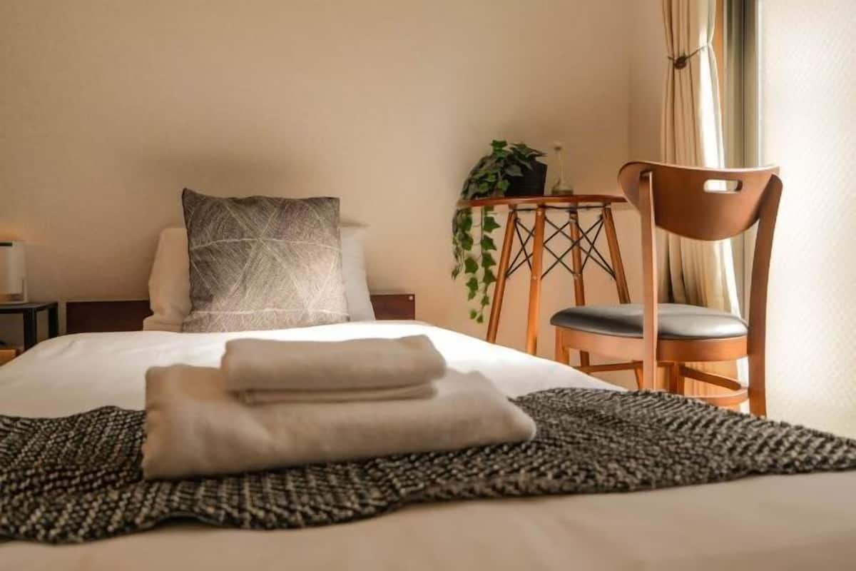Una cama bien hecha con una toalla doblada, una almohada estampada, una mesita con una planta y una silla.