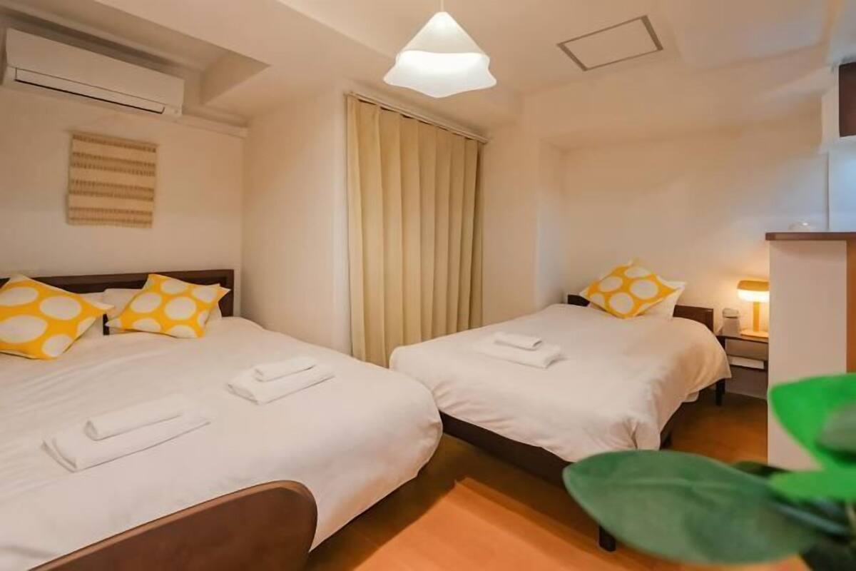 Habitación de hotel con dos camas, una mesita de noche con lámpara, una ventana con cortinas y una planta.