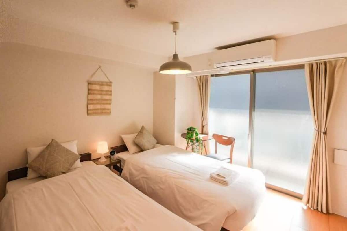 Habitación de hotel con dos camas, una mesita de noche con lámpara, una planta colgante y vistas al mar a través de un ventanal grande.