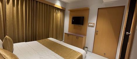 A hotel room with a bed, curtains, a television mounted on the wall, a wooden cabinet, and a door.