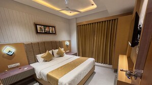 A modern hotel room with a large bed, a bedside table with a lamp, a wall-mounted artwork, and a window with curtains. - Hotel Prenix Sky- Gota (Ahmedabad)