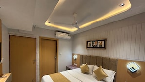 A modern hotel room with a large bed, a headboard with built-in lighting, a ceiling fan, and a wall-mounted air conditioner. - Hotel Prenix Sky- Gota (Ahmedabad)