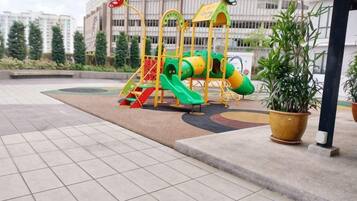 A playground with slides and climbing structures, surrounded by potted plants and a paved area.