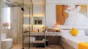 A modern hotel room with a glass-enclosed shower, a bed with white bedding and a yellow pillow, a painting on the wall, and a bathroom with a toilet, sink, and towel rack. - Cubicity Ton That Tung (Ho Chi Minh City)