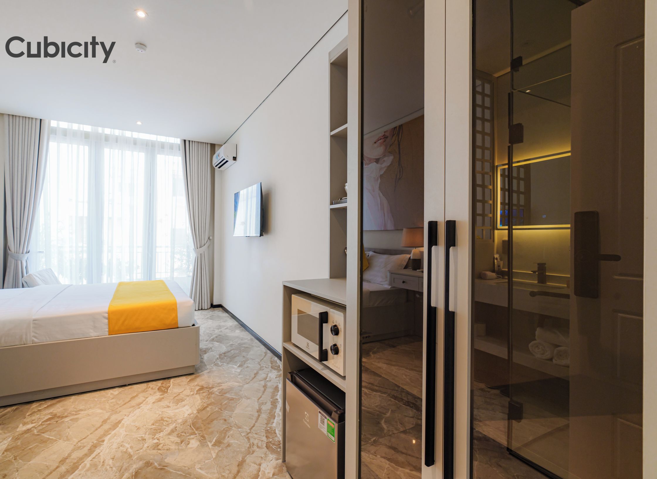 Una habitación de hotel moderna con una cama, microondas integrado, mini refrigerador y baño con paredes de vidrio.