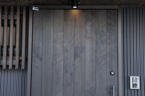 A large, dark wooden door with a vertical panel design, a small light fixture above it, and a metal panel to the right. - Trive Kamejima (Nagoya)