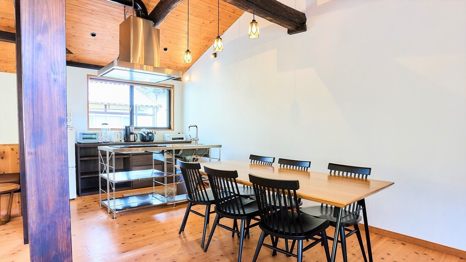 Un comedor con una mesa y sillas de madera, una cocina al fondo y un ventanal que proporciona luz natural.