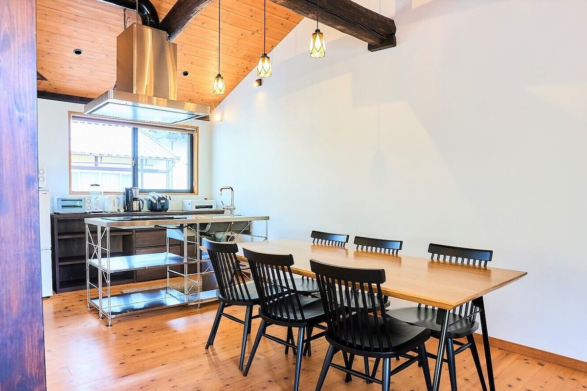 A dining area with a wooden table and chairs, a kitchen in the background, and a window providing natural light.