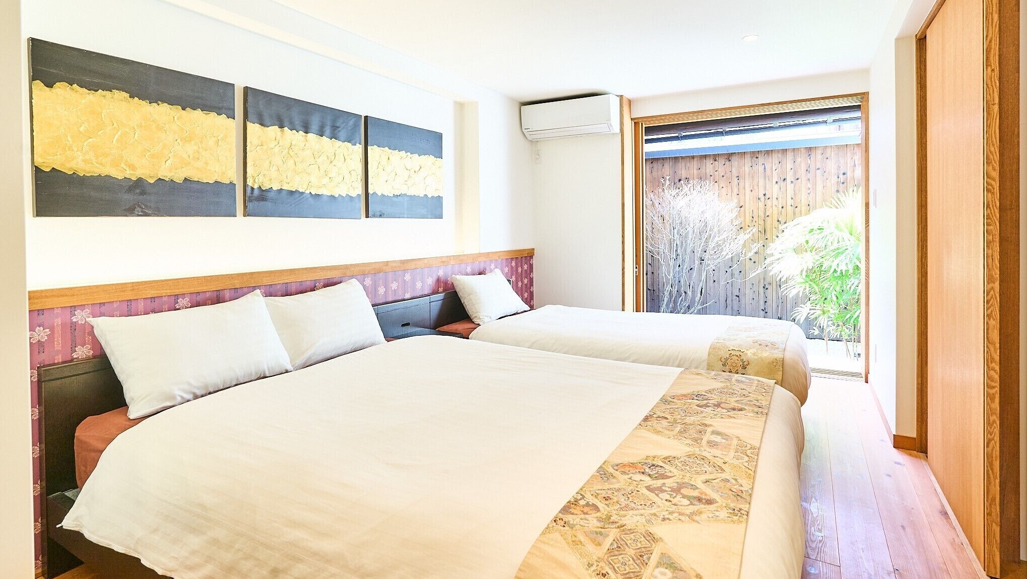 A hotel room with two beds, a large abstract painting, an air conditioning unit, and a view of greenery through a wooden door.