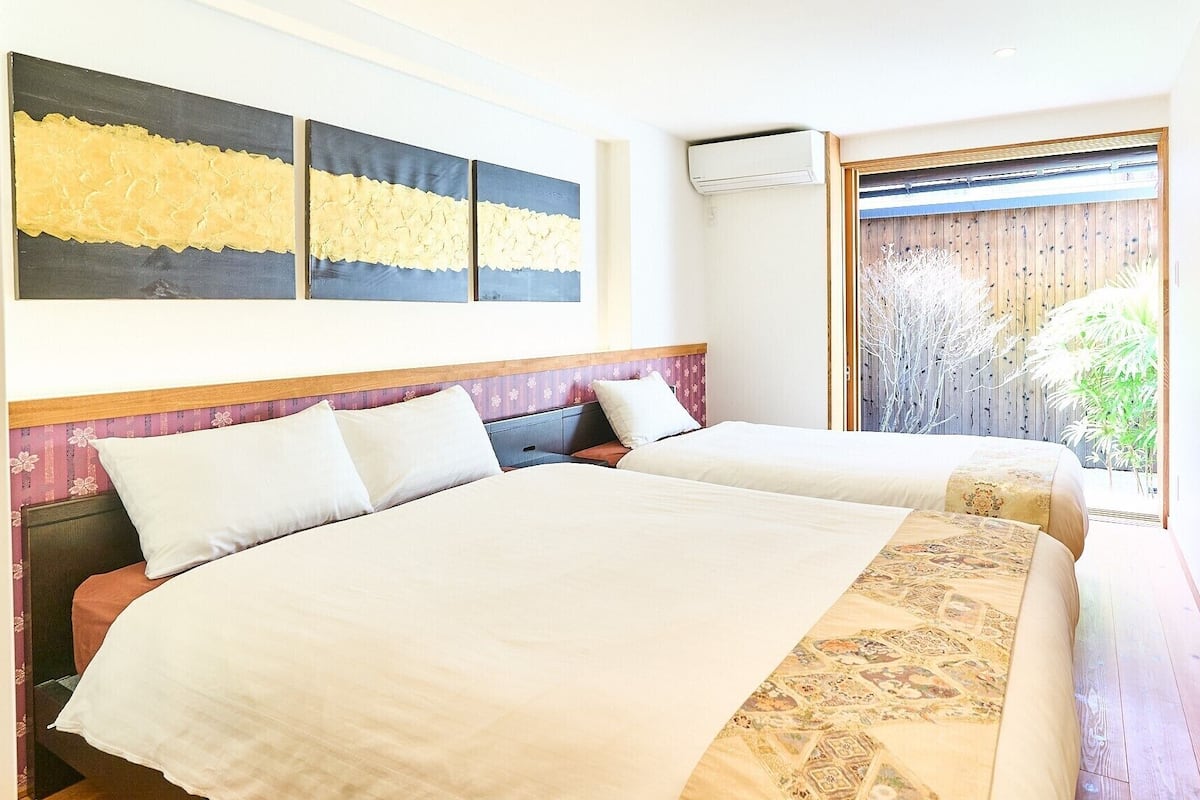 A hotel room with two beds, a large abstract painting, an air conditioning unit, and a view of greenery through a wooden door.