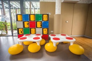 Una habitación con un ventanal grande, un estante en la pared con bloques de colores y varios taburetes amarillos con detalles rojos.
