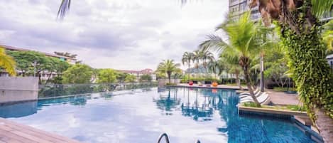 Una piscina con cubierta de madera, rodeada de palmeras y follaje, bajo un cielo despejado.