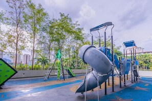 Een speeltuin met glijbanen en klimconstructies, omringd door bomen en een stadsgezicht op de achtergrond.