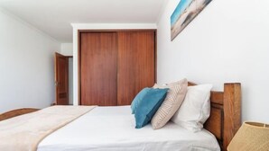 Een slaapkamer met een houten garderobe, een bed met witte beddengoed, een blauw kussen, een beige kussen en een houten nachtkastje met een lamp.