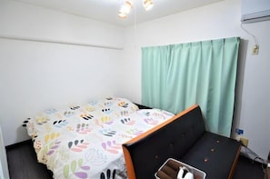 A small, well-lit room with a bed, a chair, and a window with curtains. - Comfort CUBE PHOENIX Miyazaki (Miyazaki)