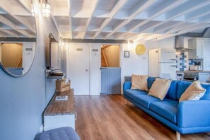A modern living room with a blue sofa, wooden flooring, a kitchen area with white cabinets, and a staircase leading to another level. - Beachwood - 2 Bedroom Cottage - Penally (Tenby)