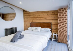 Habitación de hotel con pared de madera, un espejo redondo, una cama con ropa de cama blanca, un taburete pequeño y un radiador.