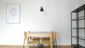 A dining area with a table and chairs, a black pendant light, a framed abstract artwork, and a metal shelving unit. - RentPlanet - Apartament Brneńska (Poznań)